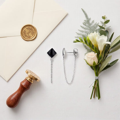 Boucles d’oreilles à losange noir et chaîne argentée disposées sur fond clair près de fleurs blanches et d’une enveloppe cachetée - KENVIA