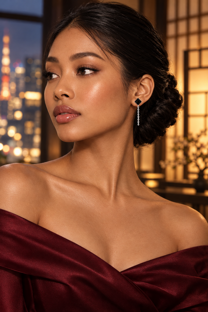 Boucle d’oreille à losange noir et mini chaîne argentée portée par une femme en robe bordeaux à épaules dégagées.